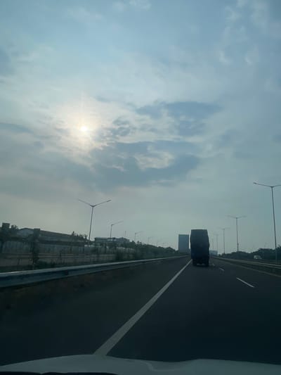 The image is a vertical, eye-level, medium shot taken from the perspective of someone inside a vehicle traveling on a highway in Kosambi Barat, Indonesia. The sky is overcast with a bright sun peeking through the clouds in the upper left quadrant. The highway has multiple lanes, with a white line marking the lanes. A dark-colored truck is traveling in the right lane ahead of the viewer's vehicle. Along the left side of the highway, there is a guardrail, followed by a strip of vegetation and then buildings. Streetlights are visible along both sides of the highway. The overall lighting suggests it is daytime, likely mid-morning or late afternoon due to the angle of the sun.