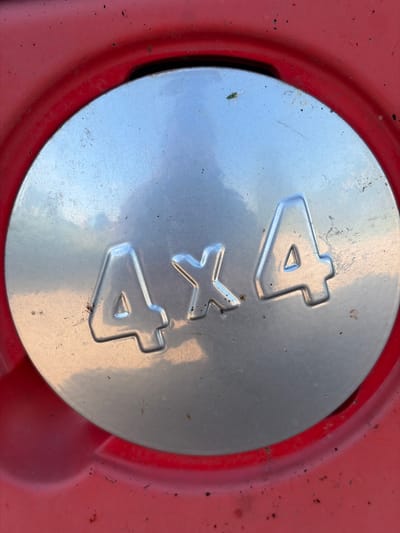 The image is a close-up, top-down view of a metallic circular emblem with "4X4" embossed on it, set within a red textured surface. The emblem is silver and shows signs of wear and minor rust spots. A small green insect is visible on the upper left quadrant of the emblem. The red surface surrounding the emblem appears to be part of a vehicle's exterior, possibly a car or truck, and exhibits a matte finish with some dirt specks. The overall lighting suggests an outdoor environment. The exact location in Zarech'e, Russia cannot be determined from the image alone.