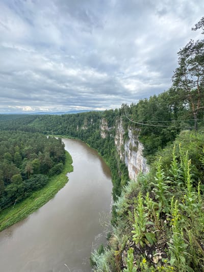 The image depicts a wide, muddy river flowing through a lush, green valley. The riverbanks are lined with dense forests, with steep, rocky cliffs rising on the right side of the frame. A suspension bridge spans the cliffs, with two figures visible crossing it. The sky is overcast with dramatic clouds. The scene is set in the city of Kul'metovo, Russia, known for its natural beauty and outdoor recreational opportunities.