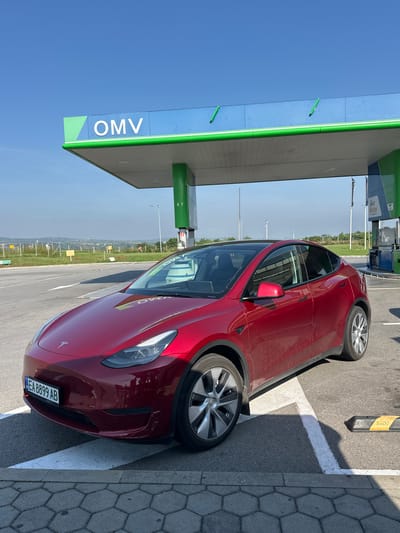 A metallic dark red Tesla, identified by its visible logo and body style (consistent with a Model Y), is parked in a designated bay at an OMV service station in Rogozinovo, Bulgaria. The vehicle displays a Bulgarian license plate, 'EA 8899 AB', mounted on its front bumper. Minor scuffs are present on the lower right side of the front bumper, and the car's exterior shows a layer of dust, particularly on the lower panels. Reflections of the blue OMV canopy and its white lettering are discernible on the car's hood and windshield.
The OMV station features a prominent blue canopy with the white "OMV" logo, accented with green trim and support pillars. Gas pumps are visible further back under the canopy. The parking area consists of asphalt pavement with white painted lines and a section of hexagonal gray pavers bordering the foreground. A black and yellow parking stop barrier, marked "STOP", is positioned to the right of the Tesla. The scene is illuminated by natural daylight under a clear blue sky. In the distant background, a landscape of undulating hills is visible, partially obscured by a slight atmospheric haze, with a fence and various signs situated along the roadside. No individuals are visible within the frame.