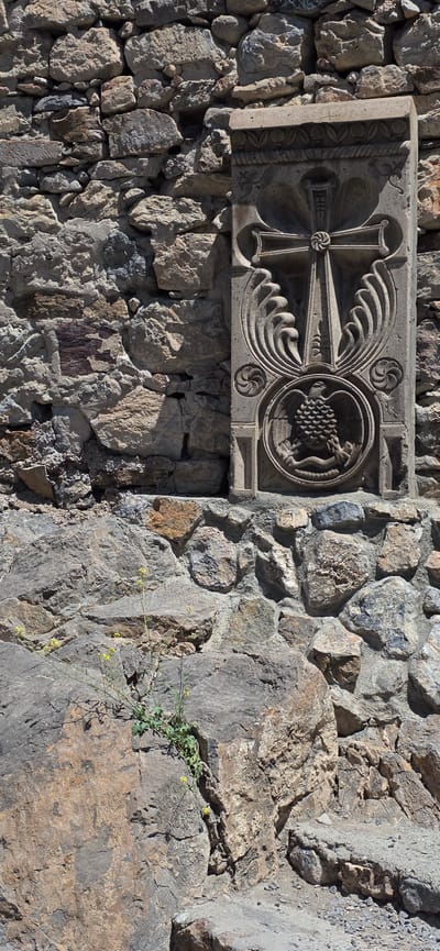 In Lusarat, Armenia, a stone carving is embedded in a rough, multi-colored stone wall. The carving is rectangular and vertically oriented, featuring intricate details. At the top, there is a decorative border with leaf-like motifs and a rope pattern. Below this, a large cross is centrally depicted, with ornate, wing-like elements extending outwards from its sides. A circular emblem at the bottom of the carving contains a detailed relief of an eagle perched on a snake. The surrounding stone wall is constructed from irregular, weathered stones of varying sizes and hues, ranging from light gray to brown and reddish-brown. A small cluster of green plants with tiny yellow flowers is growing from a crevice in the lower part of the wall, adding a touch of nature to the ancient stonework. The scene is bathed in bright sunlight, casting shadows that highlight the textures of the stone and the relief carving.