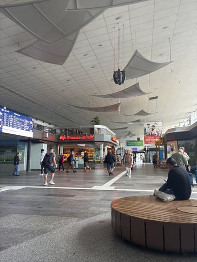 The image shows the interior of what appears to be a transit hub or train station, potentially located in Cottbus - Chóśebuz, Germany, based on the signs and language. The high, white ceiling is adorned with numerous suspended, sail-like fabric structures. A large, round, wooden bench occupies the right side of the frame, with a person seated on it and several people visible behind the bench with their luggage.

Along the right side of the image are shops or food vendors, including a "bap Sternenbäck" cafe, indicated by a sign. A digital departure board displaying train schedules is visible on the left. Several individuals are walking or standing in the central walkway. Various advertisements and signage are displayed on the walls and above the shops. The floor consists of gray tiles, and the overall lighting is bright and diffuse.