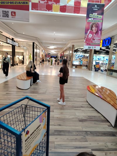 The image depicts the interior of a shopping mall in Wołomin, Poland. In the foreground, a blue shopping cart with a sign attached to it is visible. The sign displays text in Polish and an illustration of a cartoon character wearing a face mask. In the middle ground, two individuals are seated on a curved wooden bench. One has long blonde hair and is wearing dark clothing, while the other is obscured from view. Another individual, wearing a light green shirt and dark pants, walks away from the camera towards a store labeled "RESERVED." In the background, several shops are visible, including "RESERVED" and "AGD," which sells home appliances. Banners hang from the ceiling, one featuring an advertisement for "GALERIA WOŁOMIN" with the slogan "TY KUPUJESZ, MY NAGRADZAMY!" (You buy, we reward!). The mall has a modern design with a tiled floor and a suspended ceiling.