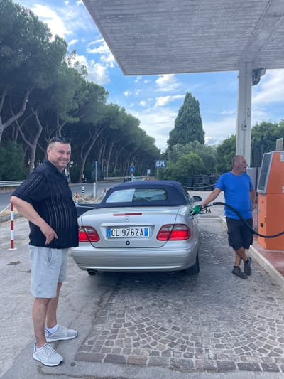 The image shows two men at a gas station in Puntone di Scarlino, Italy. One man, wearing a blue t-shirt and black shorts, is holding a green gas pump nozzle and fueling a silver convertible car. The other man, wearing a black short-sleeved shirt and light grey shorts, is standing next to the car, looking at the camera with a slight smile. He is wearing white athletic shoes. The car has an Italian license plate that reads "CL 976ZA". The gas station has an orange pump and a grey roof structure. The ground is paved with cobblestones. In the background, there are trees, a cloudy sky, and a sign with "GPL" on it. The scene appears to be taking place during the daytime, likely in the afternoon, with fair weather.
