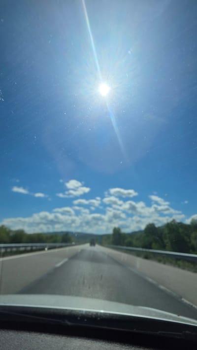 The image shows a view from inside a vehicle traveling on a highway. The scene includes a clear blue sky with the sun shining brightly and some scattered white clouds. Alongside the highway, there are trees and greenery. The location appears to be a road in Blajievo, Bulgaria.