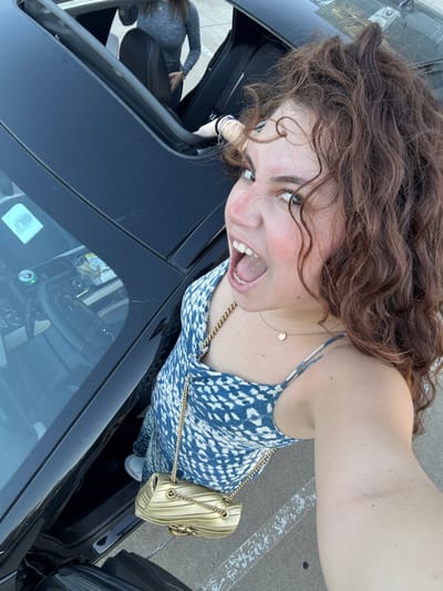A woman with curly brown hair is shown from an upper angle, leaning into a black car. She wears a patterned blue and white dress with thin straps and a gold chain bag. Her mouth is open in an expression. Inside the car, another person sits in the driver's seat, visible through the open roof. A drink can is seen on the car's dashboard through the windshield.