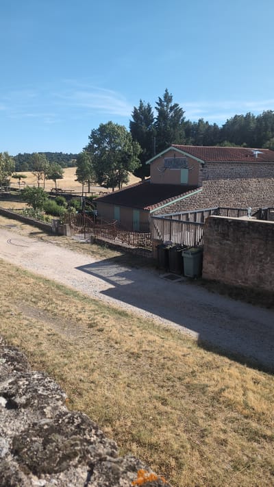 The image depicts a rural scene in Chambles, France, characterized by dry, golden grass in the foreground and middle ground. A dirt road curves through the scene, with a shadow cast by an unseen structure creating a striped pattern on it. To the right, a stone wall encloses a building complex. One building has a terracotta tiled roof and a cream-colored facade with a sign that reads "Les FOLEI". Next to it, another building features a wooden fence and several dark green and one light green trash bins. In the background, rolling hills covered with trees and fields under a clear blue sky suggest a warm, sunny day. The overall impression is one of a tranquil, outdoor location.
