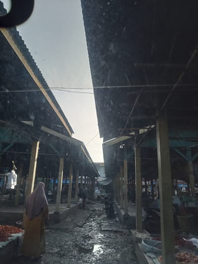The image depicts an outdoor market scene in Ujung Tanjung, Indonesia. Two covered stalls with corrugated metal roofs, supported by wooden pillars, face each other, creating an aisle. The ground is muddy and uneven, with puddles of water, suggesting recent rain. Stalls are lined with various goods, including piles of tomatoes and other produce. In the left stall, a person wearing a hijab and a yellow garment stands facing away from the camera, near a display of tomatoes. Several motorcycles are parked in the aisle. The overall lighting suggests it is either early morning or late afternoon.