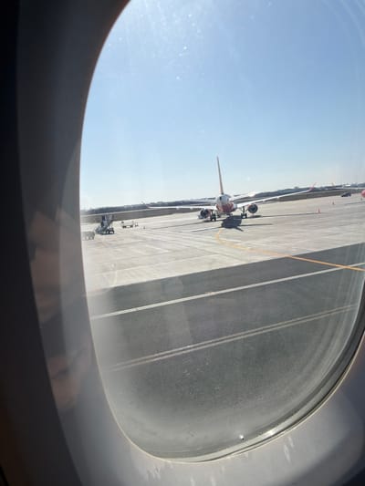 The image depicts an airport scene observed from inside an aircraft through a window, located at Henri Coandă International Airport near Tunari, Romania. A prominent passenger aircraft, displaying a white fuselage with red/orange markings on its tail fin and engine nacelles, is parked on a concrete apron, viewed from its rear. The aircraft casts a shadow directly beneath it, indicative of a high sun angle. To the left of the primary aircraft, ground service equipment, including mobile stairs and baggage carts, is visible. The apron features distinct gray and darker gray sections with yellow and white painted lines. The background reveals a distant strip of dark foliage against a clear, bright blue sky, with sun glare visible on the aircraft window. No specific individuals or actions are discernible.