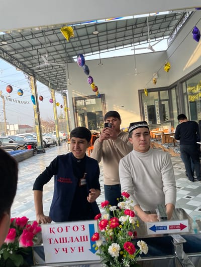 The scene depicts a semi-enclosed outdoor dining or gathering area in Chinabad, Uzbekistan. Four male individuals are visible in the foreground and midground, with a fifth partially visible in the background.
In the immediate foreground, to the left, the back of a head with dark hair is partially visible. To its right, a young male with dark hair and a smiling expression stands behind a counter. He wears a dark blue vest over a black long-sleeved shirt, with a red ID badge visible on his chest (text appears reversed due to reflection, possibly stating "YUDNIA MADH E O" or similar, and "ТУРКИЯ МАХСУЛОТЛАРИ" on the vest). On the counter in front of him are artificial pink flowers and a white sign with blue Uzbek text "КОГОЗ ТАШЛАШ УЧУН" (FOR PAPER DISPOSAL) and a downward arrow.
To his right, a second young male with dark hair wears a white turtleneck sweater and a black and white patterned Uzbek "do'ppi" cap. He stands behind a handwashing station featuring a metallic faucet. Small signs on the faucet indicate "СОВУК" (COLD) and "ИССИК" (HOT). Artificial red and white flowers are arranged on the counter to his left. He looks directly forward.
In the midground, between the two foreground individuals, a third young male with dark hair wears a light beige long-sleeved polo shirt and blue jeans. He holds a black smartphone horizontally, appearing to photograph or video-record the scene, looking at the phone's screen.
In the background, to the right, a fourth male wearing a dark long-sleeved shirt and dark pants is partially visible, facing away from the camera, interacting with objects on a table inside a building structure.
The environment features a metal-framed roof with exposed beams, supporting several white ceiling fans. Colorful metallic foil balloons (yellow, purple, blue, gold, red, orange) and white pennant flags are strung across the ceiling. The floor is covered with patterned tiles in white and grey/blue. Through large glass windows and an open section to the left, parked vehicles (including a dark SUV and white sedans) are visible, along with utility poles and distant buildings. Inside the main building structure, wooden tables with patterned seating are visible, and banners reading "RAMADAN MUBARAK" are displayed vertically on the windows. Natural daylight illuminates the scene, casting distinct shadows.