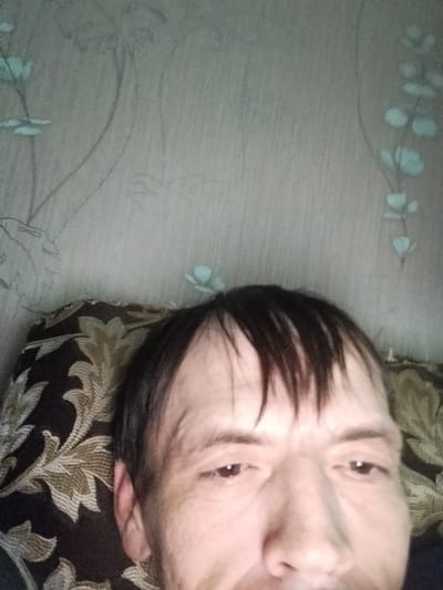 A close-up, low-angle selfie features a middle-aged Caucasian male lying down, looking directly into the camera. His face, partially obscured by a patterned pillow and dark hair that falls over his forehead, is the primary focus. The background consists of a wall with a faded floral wallpaper pattern in shades of grey and light blue. The lighting appears to be artificial and dim, casting subtle shadows on the man's face. The setting is indoors, suggesting a domestic environment. The specific location of Zyuzino, Russia, is not discernible from the visual elements present in the image.