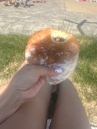 A close-up shot focuses on a person's hand holding a partially eaten pastry wrapped in clear plastic. The pastry is golden brown with a dusting of powdered sugar, and a visible chunk has been removed, revealing a dark filling. The hand, with neatly manicured fingernails painted a pearlescent purple, is positioned in the foreground. The background suggests an outdoor setting with green grass, sand, and a paved walkway where people and a bicycle are visible. The scene appears to be near Lake Sviatoz, Svitiaz, Ukraine, possibly a park or recreational area due to the presence of sand and grass. The overall lighting indicates a bright, sunny day.