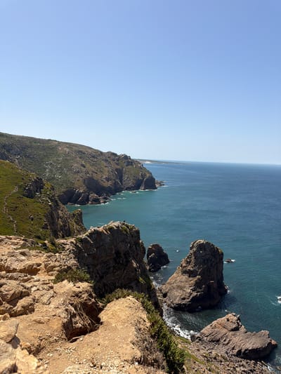 This is a vertical, medium-shot photograph of the rugged coastline near Cabo da Roca, located in the civil parish of Colares, Sintra Municipality, Portugal. The image captures a dramatic vista of steep, rocky cliffs that plunge into the deep blue Atlantic Ocean. The cliffs are covered in sparse, green vegetation, with a winding dirt path visible on the left side of the frame, indicating a hiking trail.

In the foreground, weathered, tan-colored rocks dominate the scene, with some patches of green succulent-like plants growing between them. The rocks are uneven and textured, suggesting a natural, untamed environment.

The middle ground features more imposing cliffs that jut out into the sea, with large rock formations rising from the water. The ocean is a rich, dark blue, with white foam where waves crash against the rocks. The water appears choppy in some areas.

In the background, the coastline stretches out into the distance, with more rugged terrain and hints of distant landmass. The sky is a clear, pale blue, devoid of clouds, suggesting a bright, sunny day. There are no people or man-made structures immediately visible in the frame, emphasizing the natural beauty of the landscape. The overall impression is one of wild, untamed coastal scenery.
