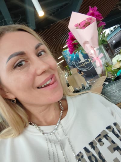 A blonde woman with defined eyebrows and a necklace is pictured in a close-up, selfie-style shot. She has her tongue sticking out, and she is wearing a white sweatshirt with a camouflage print logo. In the background, a bouquet of pink flowers is visible, along with a glass container and other floral arrangements. The scene appears to be indoors, possibly in a flower shop or a cafe, given the presence of a metal bucket containing cutlery. The lighting suggests an artificial light source, possibly a fluorescent tube. The environment has wooden slatted ceilings, and a blurred image of a sign is visible behind the flowers. There is no specific indication of the location within Amursk, Russia.