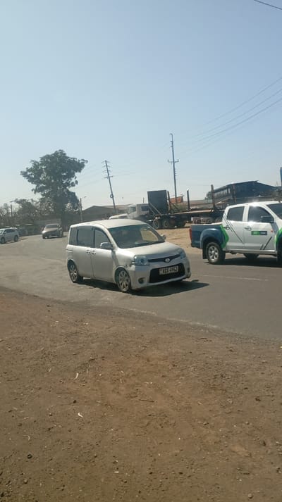 The image presents a sunlit outdoor scene, likely on a clear day, depicting a road with several vehicles. In the center foreground, a silver Toyota Sienta minivan is prominently featured, appearing to be in motion, possibly making a turn. The minivan has a visible front license plate displaying "KBS 494J," indicating its registration in Kenya. The vehicle is equipped with dark window visors.
To the right of the minivan, a white pickup truck is also on the paved road. This pickup truck is distinctive with green and blue graphical decals on its side, and the word "Safaricom" is faintly discernible within the branding, suggesting it belongs to the Kenyan telecommunications company. Further in the background, a number of large vehicles, including semi-trailer trucks with flatbed trailers, are visible, implying an industrial or commercial area adjacent to the road.
On the far left, a large, leafy green tree stands in the mid-ground. The immediate foreground on the left side of the image consists of bare, dry, brown earth or dirt, scattered with small stones and debris, suggesting an unpaved shoulder or roadside. The main thoroughfare is a dark, paved road. Above the horizon, numerous utility poles supporting a network of power lines crisscross against a clear, pale blue sky. In the further distance on the left, another dark-colored sedan is partially visible on the road, along with hints of another light-colored vehicle. The overall impression is of a bustling road in a sunny, possibly semi-urban or industrial environment.