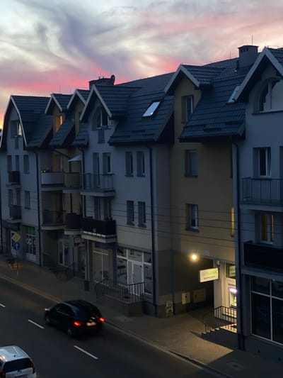 This media file depicts a street scene in Swarzędz, Poland, at dusk. The primary subjects are a multi-story building with a dark tiled roof and a black car driving on the adjacent road. The building features several windows, balconies, and a street-level storefront. Above the building, a dramatic sky with streaks of pink and purple clouds suggests the sun is setting.
The storefront area shows signs for "BANK" and "BANKOMAT," indicating a bank or ATM is present. A bright light illuminates the entrance to the bank. The road has clear white lane markings, and another vehicle, a white van, is partially visible in the lower right corner. The overall impression is of a quiet urban evening. The lighting suggests it is either early evening or late twilight, with artificial lights beginning to dominate the scene. The presence of the sky colors indicates a potentially pleasant, though rapidly darkening, weather condition. No people are clearly visible in the frame.