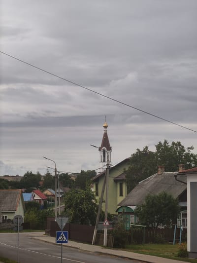 The image depicts a residential area in Ivatsevichy, Belarus, under a cloudy sky. A street with a sidewalk and road markings runs diagonally across the foreground. A pedestrian crossing sign is visible on the right side of the street, indicating a crosswalk.
Several houses of varying architectural styles and colors are scattered throughout the scene. One prominent structure is a church with a distinctive golden dome and a steeple, partially obscured by trees. The surrounding vegetation includes lush green trees and bushes. Power lines crisscross the sky, a common element in urban landscapes. The overall impression is that of a quiet, suburban neighborhood.