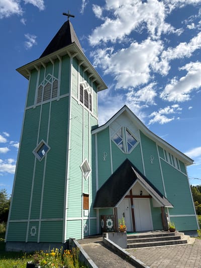 This is a low-angle, eye-level, exterior, full shot of a light green wooden church with white trim, set against a bright blue sky with scattered white clouds. The church has a tall, square bell tower with a dark pitched roof topped by a cross. The tower features louvered windows and diamond-shaped windows. Adjacent to the tower is the main body of the church, which also has a pitched roof. A covered entrance with double white doors is situated on a set of concrete stairs. Decorative white symbols, resembling the Chi-Rho, are visible on the church's exterior walls. In the foreground, there are steps leading to the entrance and a paved walkway. Some vegetation, including flowers and grass, is visible around the base of the church and on the left side of the image. The scene appears to be located in Läskelä, Russia. There are no people visible in the image.