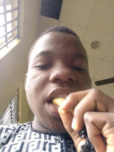 In the city of Eleweran, Nigeria, a young person is captured in a close-up, low-angle selfie. They are in the process of eating a piece of food, holding it with their right hand, which is positioned near their mouth. The person has dark skin and short dark hair. They are wearing a black and white patterned shirt. The background shows a ceiling with light fixtures and a window with a metal grate. The lighting suggests it is daytime.
