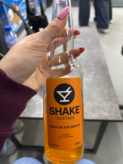 A person's hand with pink painted fingernails is holding a clear glass bottle with an orange label. The label has black text that reads "SHAKE COCKTAILS" and "SEXX ON THE BEACH." Below this, it says "low alcohol carbonated cocktail" and "330ml" and "alc. 5% vol." The background is blurred but appears to be an indoor setting with people walking in the distance. The image does not contain any information about the city of Satovcha, Bulgaria.