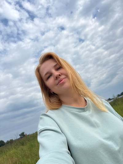 A woman with blonde hair is taking a selfie outdoors. She is wearing a light blue sweatshirt. The background shows a grassy field under a cloudy sky. The location is Breytovo, Russia.