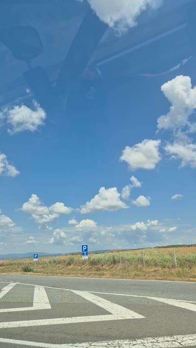 The image depicts a roadside scene with a bright blue sky filled with scattered white clouds. In the foreground, a gray asphalt road with white lane markings is visible. Beyond the road, a dry, grassy embankment leads up to a fenced field with sparse vegetation and some flowering plants. Two blue and white parking signs are placed along the roadside. The sign on the right appears to have additional text below the main "P" symbol. In the distance, rolling hills are visible under the expansive sky. Reflections of the car's interior, including what appears to be a rearview mirror and part of the dashboard, are visible on the windshield, indicating the photo was taken from inside a vehicle. The location is identified as Zheleznik, Bulgaria.