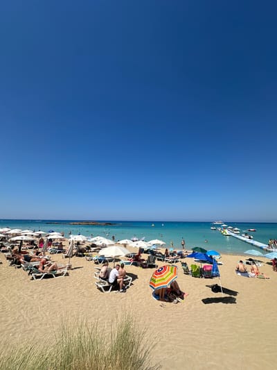 This is a wide-angle, eye-level shot of a crowded beach in Protaras, Cyprus, on a clear, sunny day. The foreground is dominated by sand, with a few tufts of dry grass on the left. The middle ground depicts a busy beach scene with numerous people sunbathing on lounge chairs, sitting under umbrellas, and walking around. White beach umbrellas are prevalent, along with a few colorful ones. The background features the turquoise Mediterranean Sea, dotted with several boats and a floating water park with a long pier. The sky is a vibrant, cloudless blue, occupying the upper half of the image.