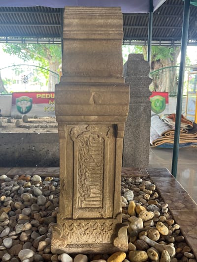 A stone monument, likely a tombstone, stands prominently in the foreground, adorned with intricate carvings. It is surrounded by a bed of smooth, rounded stones of various sizes and colors. The monument is situated outdoors, under a covered structure with a sloped, ribbed roof. In the background, other similar stone markers are visible, suggesting a historical burial site. Banners with logos and text are displayed behind the monuments. Lush green trees and foliage can be seen beyond the covered area. The overall environment appears to be a peaceful, historical site in Beuringen, Indonesia, possibly a cemetery or a sacred place of remembrance.