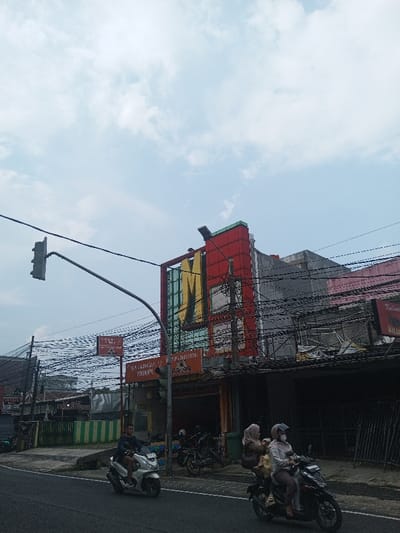 The image captures a daytime street scene in an urban or semi-urban environment, likely under an overcast sky. The upper half of the frame is dominated by a light blue sky with scattered white, wispy clouds.
In the mid-ground, a prominent multi-story building stands out. Its upper facade is painted a vibrant red with a green trim along the top edge. A large, stylized yellow letter "M" is fixed to the red facade, appearing to span at least two stories. The vertical bars within the "M" feature a light blue or greenish hue. Numerous black utility wires crisscross densely in front of and around this building, connecting to poles and other structures.
Below the large "M" building, at street level, there are shopfronts. One shop has a red sign where the word "KULINER" is clearly visible on the right side. Below this, smaller text, possibly "AYAM GEPREK" and "NASI GORENG," is present but difficult to read due to blurriness. To the left of this, another reddish-brown sign is visible, displaying text that includes "PINDANG TULANG," "PEMPEK," and "MAMA NENENG."
On the far left, a black traffic light is suspended from a long, curved gray metal pole that extends over the road. Other buildings and some foliage are visible further in the background to the left.
In the foreground, on a paved road, two motorcycles are in motion. On the left, a person wearing a dark top is riding a light-colored (possibly white or silver) scooter. On the right, a darker-colored scooter carries two individuals. The driver wears a helmet and a light-colored jacket, while the passenger behind wears a hijab and a dark top. A small child appears to be seated in front of the driver, partially obscured.
The overall scene suggests a busy commercial area, characterized by local shops, dense utility infrastructure, and common use of motorcycles for transportation.