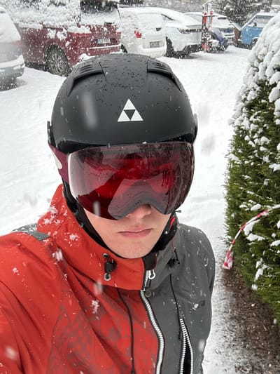 An individual is depicted in a close-up, selfie-style photograph, standing outdoors during a snowfall in Bad Hofgastein, Austria. The person is wearing a black ski helmet with a white Fischer logo (composed of three interconnected triangles) and red-tinted ski goggles. A two-tone ski jacket is visible, red on the left side and dark grey/black on the right, with prominent zippers and drawstrings. Snowflakes are actively falling and have accumulated on the helmet and jacket. The lower part of the person's face, from the nose to the chin, is visible beneath the goggles.
In the background, a parking area or roadside is covered in fresh snow. Several vehicles, heavily dusted with snow, are parked, including a red minivan or SUV with a partially visible Czech license plate ("ELY 93E") and a white SUV. Some vehicles have skis attached to their roof racks. To the right of the individual, a snow-laden coniferous tree stands, from which a piece of red and white striped barrier or caution tape hangs, partially obscured by the snow. The ground is uniformly covered in snow, indicating a significant winter event.