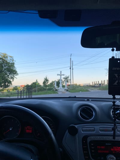 The image is taken from the interior of a car looking out through the windshield. In the foreground, the dashboard and steering wheel are visible, along with a car stereo and an air freshener hanging from the rearview mirror. Outside the car, a road leads towards a white monument with a cross and a statue. Beyond the monument, there are trees, utility poles, and power lines under a clear sky. In the distance, there appears to be a construction site with columns. The time displayed on the car's dashboard is 20:17, with a trip distance of 44.1 km and an odometer reading of 296841 km. The location is identified as Negrești, Moldova.