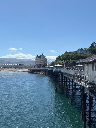 The image captures a sunny day in Llandudno, United Kingdom. A pier stretches into the sea, supported by metal framework. People can be seen walking on the pier. The shoreline features buildings and a distant mountain range under a clear blue sky with scattered clouds. The sea is shimmering with sunlight.
