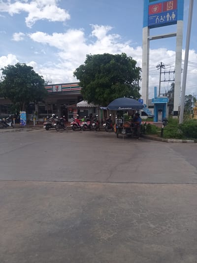 This image depicts a scene in Tha Ruea, Thailand, specifically at a PTT Station. In the background, a 7-Eleven convenience store is visible, alongside parked motorcycles. An ATM booth is present near a tall, blue and white PTT Station sign. A street vendor operates a food cart under a blue umbrella bearing the "PTT station" logo. A person is seen at the cart, presumably cooking or serving food. The foreground shows a large expanse of concrete paving, and the sky is partly cloudy.