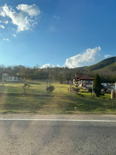 The image depicts a rural scene likely taken from a moving vehicle on a road in or near Osetenovo, Bulgaria. To the right is a two-lane road with one visible white line marking separation. Beyond a grassy verge, a chain-link fence encloses a large green field with scattered trees and wooden structures. A single-story house with a red-tiled roof, satellite dish, and garden is visible further up the slope, nestled against a tree-covered hillside. The sky above is clear blue with scattered white clouds. The scene is generally peaceful and indicates a landscape with residential and natural elements, consistent with rural areas.