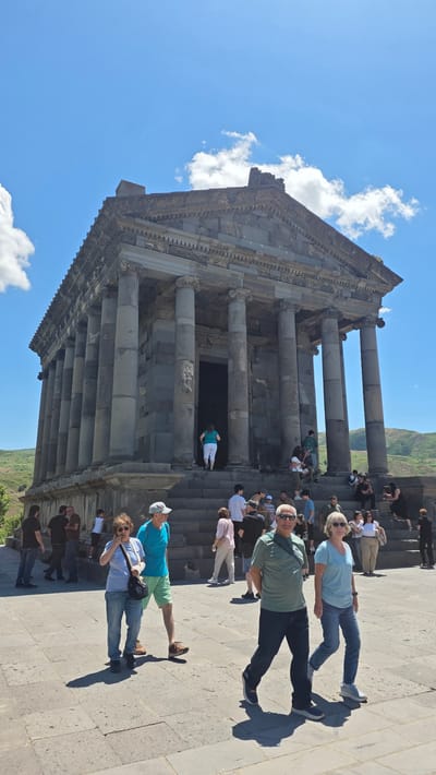 The image depicts the Garni Temple, an ancient Greco-Roman temple located in Garni, Armenia. The temple, constructed from basalt, features a prominent portico with fluted columns and a triangular pediment. Numerous people are visible in and around the temple complex. In the foreground, several individuals are walking on a paved area, while others are ascending or descending a wide set of stone stairs leading to the temple's entrance. The background reveals rolling hills and a bright blue sky with scattered clouds. The scene appears to be a sunny day, with strong shadows cast by the people and the structure.