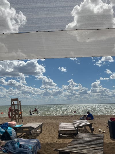 The image depicts a sun-drenched beach scene, likely in Gres-2, Kazakhstan, viewed from beneath a white mesh canopy. The sky is a vibrant blue, dotted with numerous fluffy white clouds. The sun is shining brightly, reflecting off the shimmering surface of a body of water, likely a lake or sea, in the background.
On the sandy beach, several lounge chairs are visible, some occupied. In the foreground, a pile of towels and beach gear rests on a lounge chair. Further back, a wooden lifeguard chair stands prominently, with a red lifebuoy attached. In the water, several people are engaged in various activities, including swimming and paddling on inflatable devices. Two individuals, an adult and a child, are interacting on the sand near some wooden picnic tables. The overall atmosphere is one of leisure and recreation on a warm, sunny day.