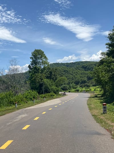 The image shows a winding asphalt road with yellow dashed lane markings, curving through a lush, green, hilly landscape under a bright blue sky with scattered white clouds. The road is bordered by a grassy verge and dense vegetation, including trees and bushes. In the foreground, there are several white posts with red tops along the side of the road. Further down the road, a signpost is visible on the right, displaying "H3" and "37". The road appears to be in a rural area, likely in the city of Ham Thuan Bac, Vietnam, given the context. The scene is serene and depicts a typical countryside road under daylight conditions. There are no visible people or vehicles in the image.