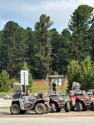 The image captures a scene at T/B Seminskii Pereval, Russia, with several ATVs parked on a dirt road in the foreground. The ATVs are various colors, including gray, blue, and red, and appear to be used for off-road excursions, as indicated by the mud on their tires and bodies. In the background, a person is standing near a wooden information board, seemingly reading the displayed content. Behind the information board and person, a dense forest of tall pine trees stretches across the landscape, under a partly cloudy sky. The environment suggests a natural, mountainous area, typical of a mountain pass.