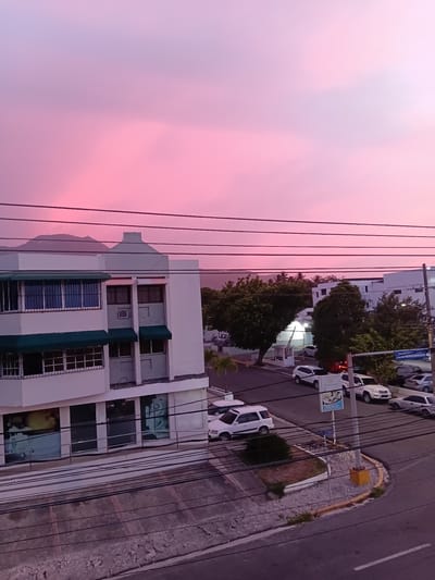 The image shows a view of a street in Puerto Plata, Dominican Republic, at dusk. The sky is a vibrant pink, characteristic of a sunset. In the foreground, there are power lines crisscrossing the frame. A multi-story white building with green awnings is visible on the left. Several cars are parked on the street and in a lot in front of a building. A street sign is visible in the middle ground. Trees and other buildings are present in the background, and a mountain range can be seen in the far distance. The scene depicts an urban environment during twilight.