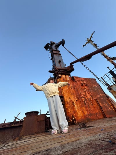 A young girl stands on the deck of a rusty shipwreck in Teriberka, Russia. She is wearing a light yellow jacket and light grey sweatpants. Her arms are spread wide as if embracing the vastness of the sky. The shipwreck, heavily weathered and covered in rust, dominates the foreground and middle ground. In the background, a clear blue sky is visible. The scene suggests a sense of exploration and wonder amidst the decaying remnants of maritime history.