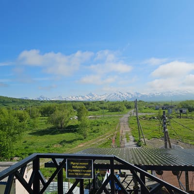 This is a vertically oriented, eye-level, outdoor photograph taken on a sunny day. The scene is a landscape with a prominent blue sky featuring white, wispy clouds on the left side. In the background, a range of mountains is visible, with snow-capped peaks contrasting against the green hillsides.

The foreground is dominated by a metal structure, possibly a balcony or viewing platform, with black metal railings. Attached to the railing is a yellow sign with black text. Below the structure, a lush green grassy area stretches towards the mountains. A dirt road or path winds through the grass, bordered by trees. Scattered across the landscape are utility poles with tangled wires. Further in the distance, there appear to be some dilapidated buildings or structures amidst the greenery.

The weather appears to be pleasant and sunny, with the clear blue sky suggesting it's daytime. The presence of snow on the mountains indicates a higher altitude or a cooler climate. The overall mood is one of natural beauty and perhaps a touch of rural isolation.

The visible text on the sign reads: "Мусор можно выбрать, спускаясь с крыши вниз по лестнице". This translates to: "Trash can be chosen, descending from the roof down the stairs." This sign likely serves as an instruction or notice related to waste disposal.

The location context is Sokoch, Russia, which helps interpret the mountainous terrain and the presence of utility infrastructure in a rural or semi-rural setting.