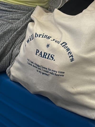 The image shows a folded white t-shirt with dark blue text on a background that includes a blue material and a gray and green striped material. The text on the t-shirt reads "will bring you flowers PARIS." Below PARIS, there is a quote that reads: "If I had a Single flower for every time I think about you, I could walk forever in my garden." The photograph appears to have been taken indoors. There are no people visible. The location is not discernible from the image.