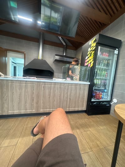 The image depicts the interior of a small food establishment, possibly a fast-food restaurant or a cafe, with a person's legs and feet in the foreground, suggesting the perspective of someone sitting. The establishment features a modern, clean aesthetic.
Key elements visible include:
* **Foreground:** A person's legs and feet are prominently displayed, wearing dark shorts and flip-flops. The floor is tiled with a wood-grain pattern.
* **Counter Area:** A counter with a wood-grain laminate finish runs across the middle ground. On top of the counter, there are various items, including what appear to be papers or menus and a small glass partition.
* **Kitchen Equipment:** Behind the counter, a large, black ventilation hood with a stainless steel flue is installed. Further back, a person, presumably an employee, is visible behind a glass display, engaged in preparing food or serving.
* **Beverage Refrigerator:** To the right of the counter, a tall, black, glass-fronted refrigerator is packed with beverages, including bottles and cans of soda and other drinks. Several yellow and pink sticky notes are affixed to the side of the refrigerator.
* **Seating Area:** A small round table with a black leg is partially visible on the far right of the image, suggesting a seating area.
* **Ceiling and Lighting:** The ceiling features exposed wooden beams and slats. Two large, flat-screen televisions are mounted on the ceiling, reflecting the overhead lighting and the exterior environment. Fluorescent lights are also visible.
* **Wall:** The walls appear to be tiled with light-colored square tiles.
The overall impression is of a functional and contemporary food service environment. The image does not provide any specific visual cues to pinpoint the exact location within Baltasi, Russia.