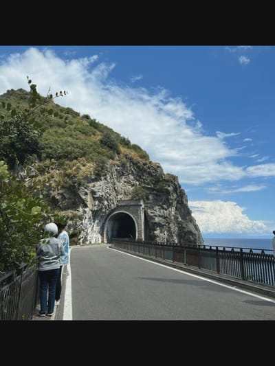 The image shows a scenic view of a coastal road with a tunnel carved into a rocky mountainside. Two people, one wearing a white helmet and a grey jacket and the other in a blue and white patterned shirt, are standing on the side of the road, looking towards the tunnel. A railing runs along the edge of the road, separating it from the steep drop to the sea on the right. The sea is visible in the background, under a bright blue sky with scattered white clouds. The rocky mountain is covered with lush green vegetation. The image does not appear to be located in Norcia, Italy, as Norcia is an inland town known for its historical architecture and seismic activity, not its coastal roads and tunnels.