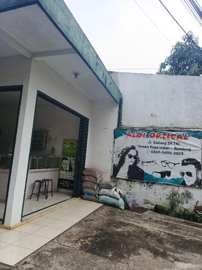 A one-story white building houses Aldi Optical. The banner displays the business name, address, and phone number along with images of people wearing sunglasses and glasses. A partially open entrance reveals the interior with a display case and optical posters. Stacked bags lean against the wall beside the doorway. The building has a concrete exterior with overgrown vegetation. Wires cut across the sky in the background.