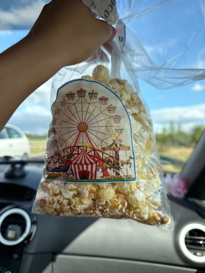 A hand is holding a clear plastic bag of popcorn in a car. The bag has a sticker with a Ferris wheel and a carousel design. In the background, a white car is visible through the car window. The scene appears to be taking place outdoors on a sunny day, with blue sky and clouds.