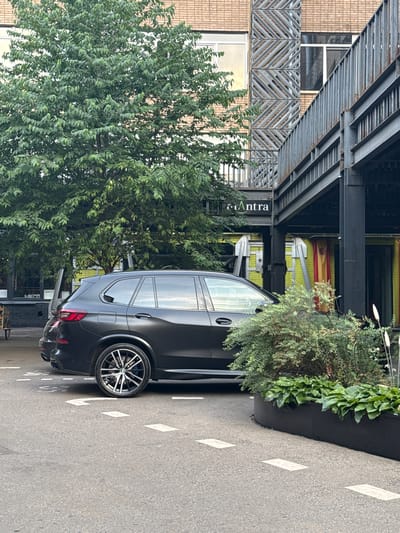 A dark-colored SUV is parked on a paved surface marked with dashed white lines. The SUV is positioned in front of a building with a metallic structure and large windows. To the left of the SUV is a large green tree with dense foliage. In the foreground, a dark planter contains green bushes and plants. The building facade is partially visible behind the vehicle, featuring a sign that reads "Antra".