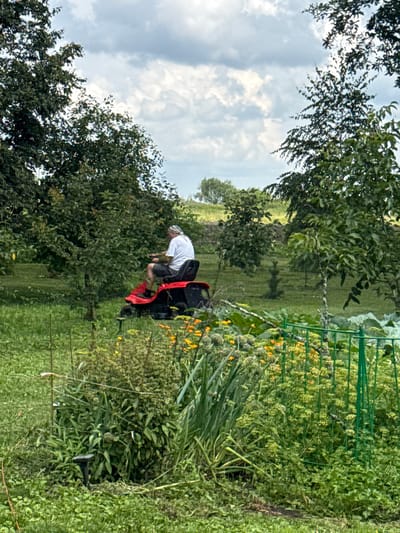 A man is riding a red and black lawnmower through a grassy yard. He is wearing a white t-shirt and dark shorts, with a bandana tied around his head. The lawnmower is in motion, and the man appears to be focused on his task. The yard is surrounded by trees, and the sky is cloudy. The overall impression is one of a rural or suburban setting. The location is the village of Bolshiye Ozerki, Russia.