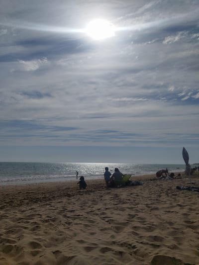 The image depicts a sunlit beach scene, likely in Rota, Spain, given the context. The sky is a mixture of blue and white clouds, with the sun prominently visible and emitting a strong glare. The sea is a shimmering blue, with gentle waves lapping at the shore. The beach itself is sandy, with rippled patterns created by the wind and tide.

In the foreground, the sand is textured and the focus of the composition. Further back, several groups of people are visible. On the left, a child is sitting on the sand. To their right, two adults are seated, one in a green chair, and a child stands beside them. In the middle ground, a group of people are lying on the sand, with one individual appearing to be tending to another. Towards the right, a tall, slender umbrella-like object stands on the beach, and further in the distance, more people are visible walking along the shoreline. The overall atmosphere is one of a leisurely day at the beach.