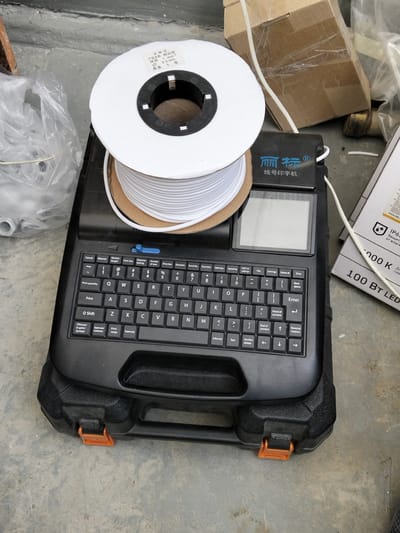 The image shows a black wire marker printer with a spool of white wire on top. The printer has a small screen and a QWERTY keyboard. The surrounding area appears to be an outdoor or industrial setting, possibly a construction site, with concrete ground and some cardboard boxes and plastic bags in the background. There are no people visible in the image. The spool of wire has a label with Chinese text, indicating "Product Name: Plume Pipe", "Specification: 1 mm² 2.5 mm", and "Quantity: 1 roll". The printer itself also has Chinese text, "丽标Ⓡ 线号印字机", which translates to "LibiaoⓇ Wire Number Printer". The overall impression is one of industrial equipment ready for use. The lighting suggests it might be daytime.